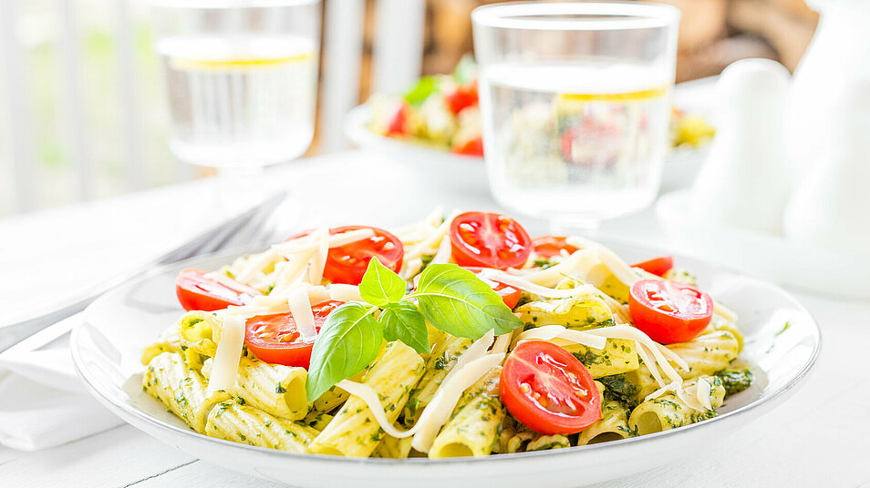 Tellergericht mit Nudeln, Tomaten, Pesto und ein Glas Wasser auf weißem Tisch