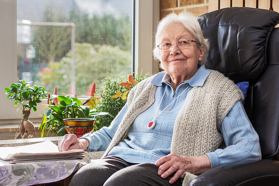 Seniorin sitzend in einem Ledersessel an einem Fenster mit einem Hausnotruf  Funkfinger um den Hals