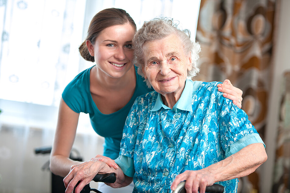 Junge Auszubildende legt fürsorglich den Arm um eine lachende alte Frau im Rollstuhl