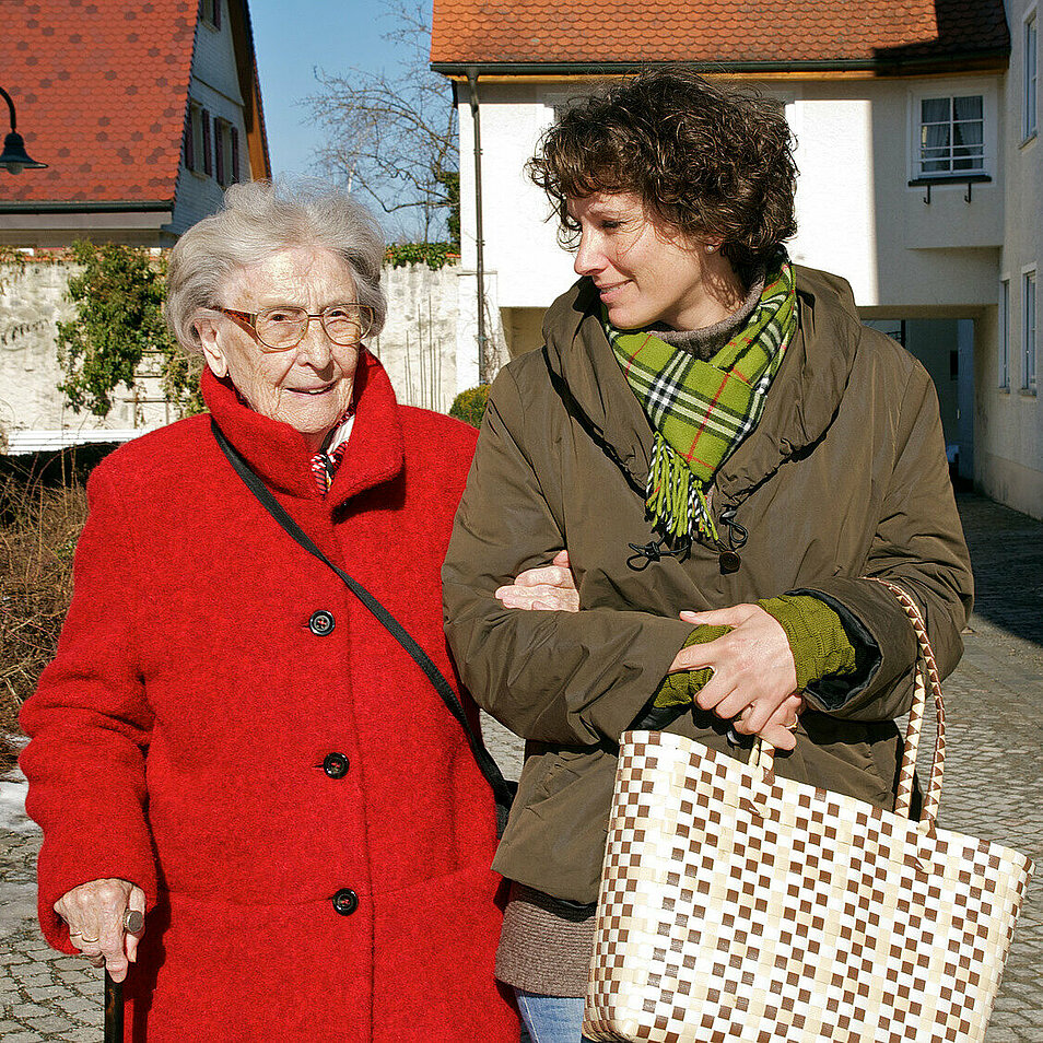 Alte Frau am Stock mit Einkaufstasche geht eingehakt bei einer Betreuerin